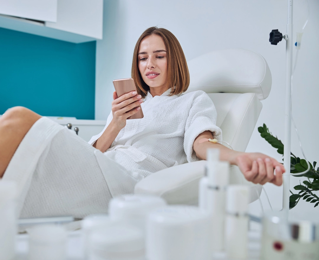 A woman in a white bathrobe relaxes in a spa chair getting IV infusion, holding a smartphone, surrounded by skincare products and a green plant